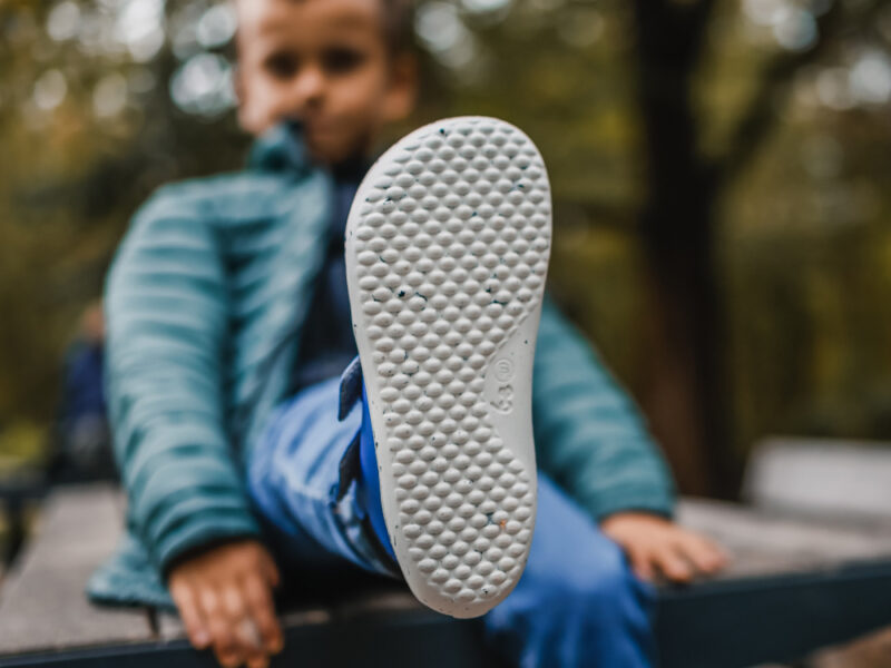 Barn på udendørs bænk viser fleksibel barfodssko med bred tåboks; minimalistisk sneaker med naturlig pasform til aktive børn.