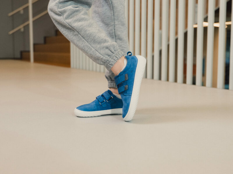 Person i grå joggingbukser bærer lyseblå barfodssko med bred tåboks på lyst gulv nær trapper og hvide gelændere.