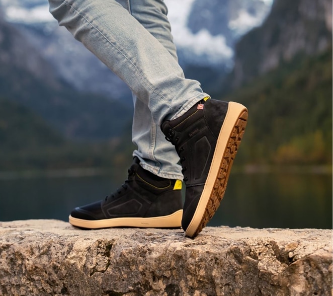 Minimalistiske sorte high-top barfodssneakers med bred tåboks, båret med lyseblå jeans udendørs ved en naturskøn bjergsø.