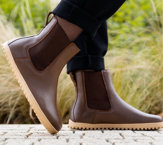 Brune barfods-chelsea boots med ribbede elastiske sider, beige såler og bred tåboks på lys mursten udendørs med græs.