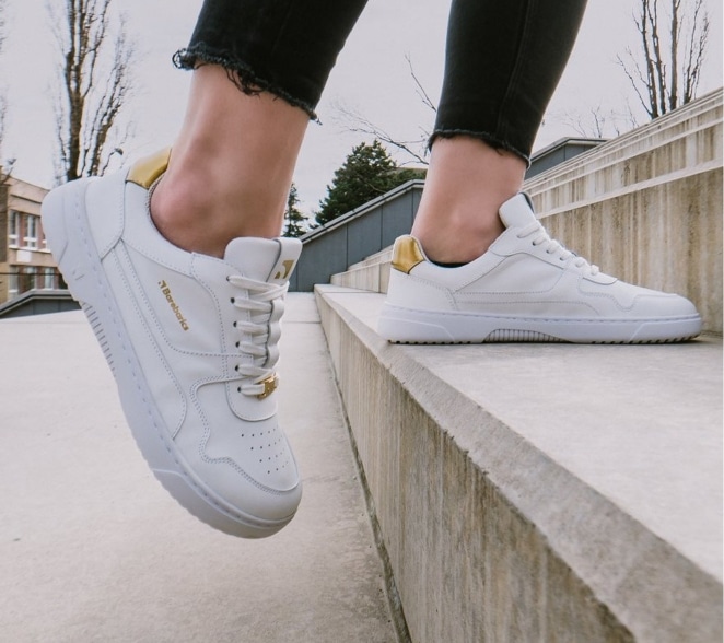 Person i sorte jeans på en betontrappe iført hvide minimalistiske barfodssneakers med gulddetaljer og bred tåboks.