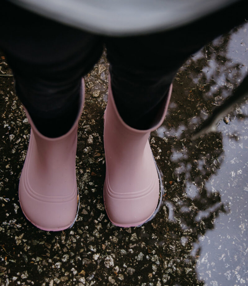 Minimalistiske pink barfodsstøvler med bred tåboks på vådt fortov, nær vandpytter, der reflekterer træer, ideelle til regnvejrsdage.